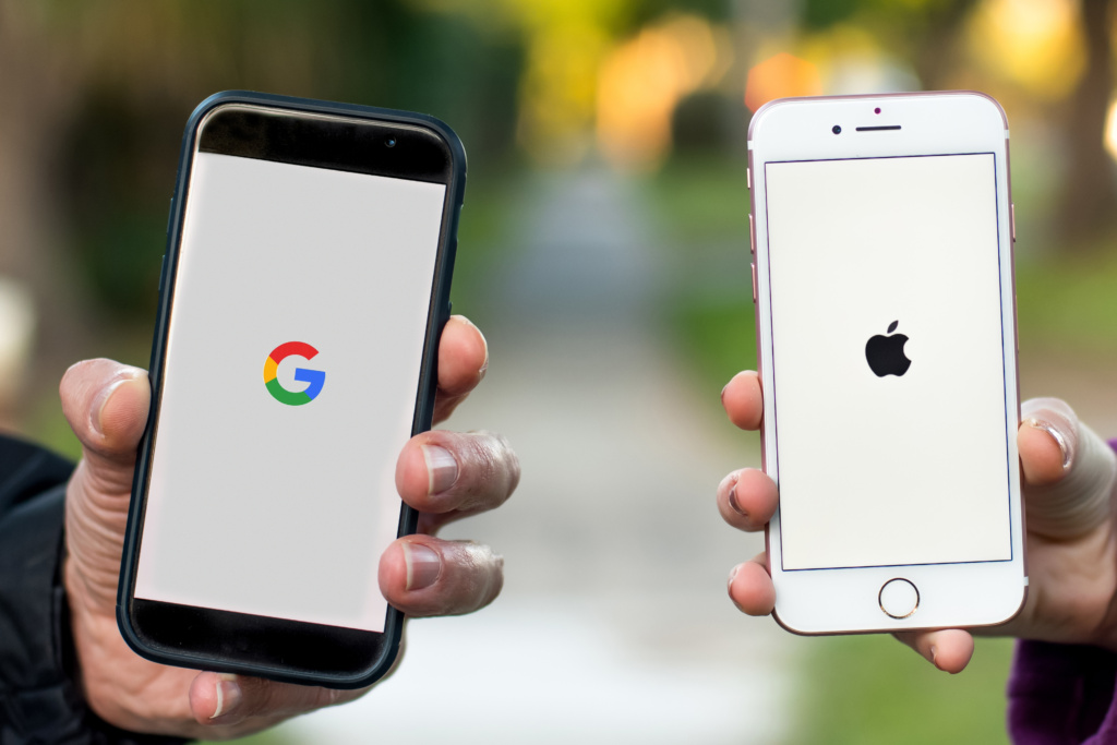 Zwei Smartphones, eines mit Google-Logo und das andere mit Apple-Logo in den Händen von Personen.